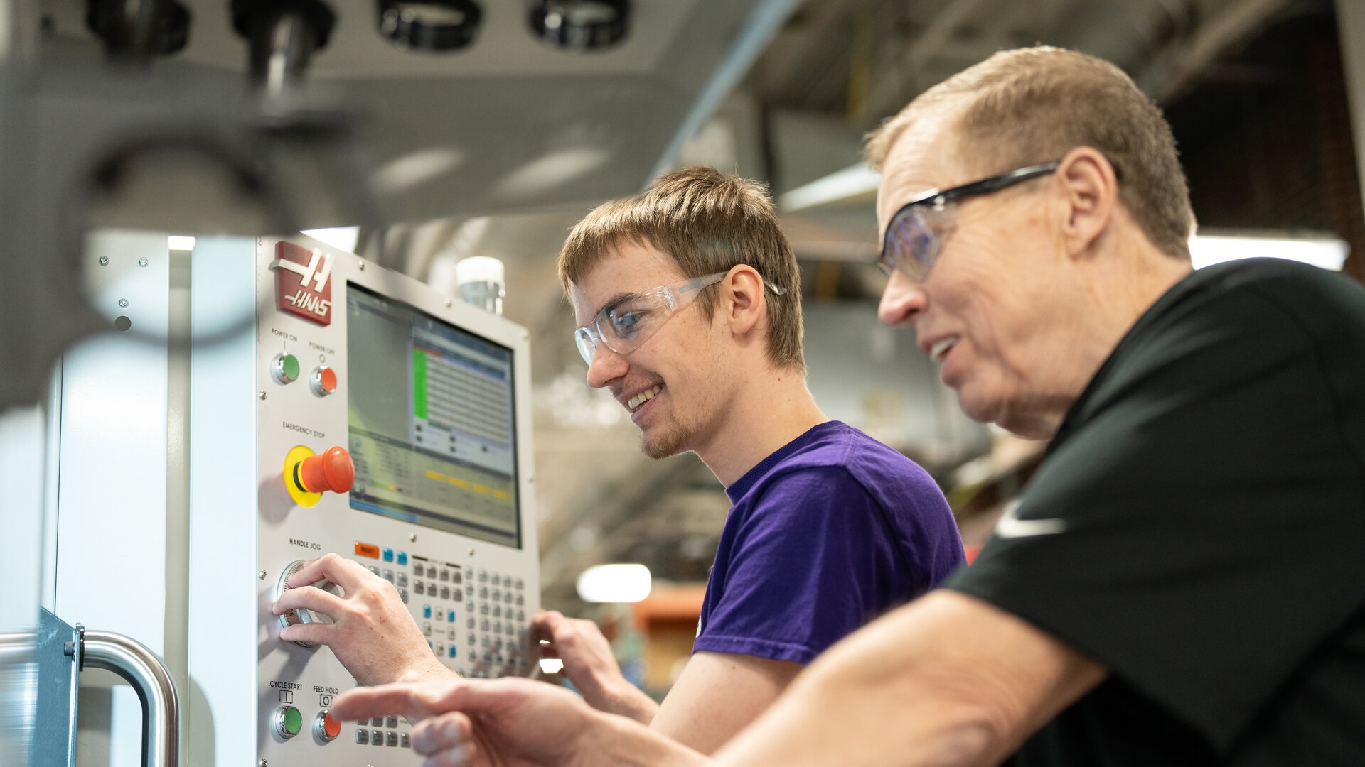 Engineering student makes adjustments on a CNC control pad while faculty assists him.