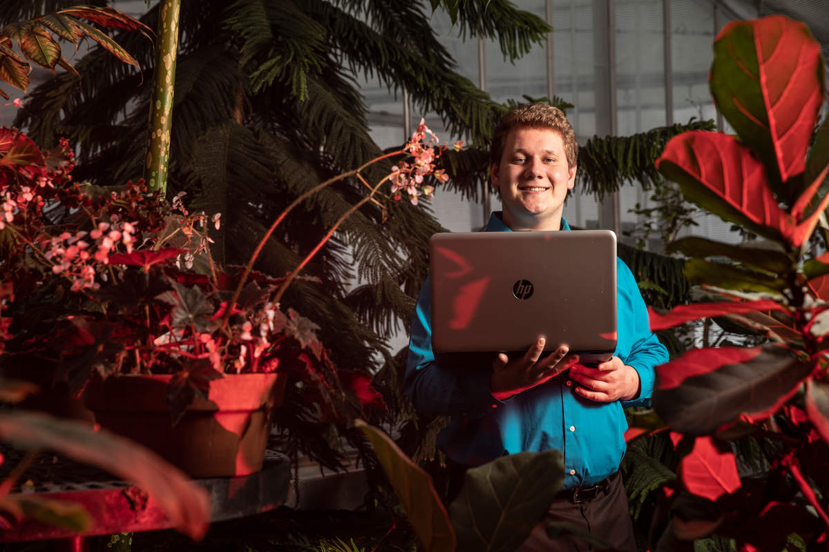 Engineering student researcher Jeremy Ziemer stands with laptop in St. Thomas greenhouse.
